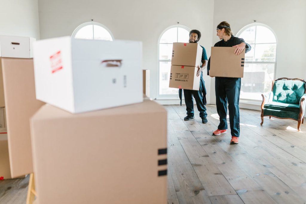 two-men-carrying-boxes-7464687 Two movers in uniform carrying boxes in a bright, new home setting.